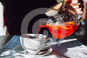 The hand of unrecognizable woman pours hot tea from glass kettle to mug sitting by table with white tablecloth.