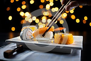 Hand using chopsticks pick Sushi and Sashimi on black background with bokeh. Fresh made Sushi set with salmon, prawns, wasabi and