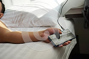 Hand using are holding smart phones. charger is plugged on the white bed, The danger of charging electricity when sleeping