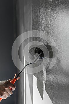 Hand using paint roller applying grey paint on white wall.