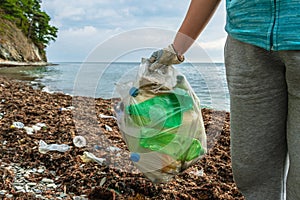 In the hand of the volunteer is a full bag of plastic bottles collected on the seashore. In the background is a polluted