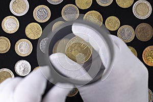 A hand in a white glove holds a coin and a magnifying glass against the background of a pattern of coins on a black cloth