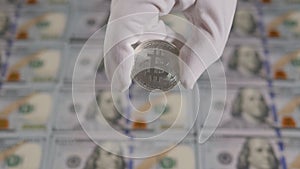 A hand in white gloves holds a silver bitcoin on the background of dollar bills.
