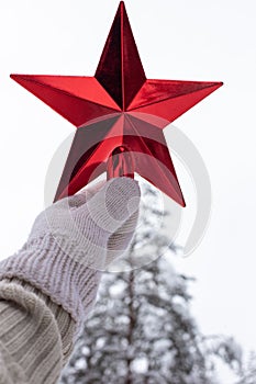 A hand in a white knitted mitten holds a red star over a snow pine tree in the forest