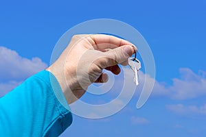 The hand of a white man holds a key on a background of a beautiful summer sky. A man in blue long sleeve holds a key