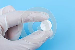 A hand in a white medical glove holds a pill close-up on a blue background. Treatment, medicine, drugs, pills, vitamins,