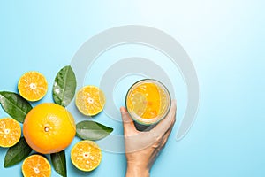 Hand of woman holding glass of orange juice and orange fruits with sliced pieces and leaves on blue background