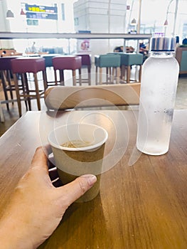 Hand of a woman holding a recyclable cardboard coffee cup. On the table is a glass water bottle, sustainable materials. Interior