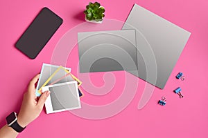 Hand of woman holding snapshot pictures, smartphone, sheets of cardboard, binder clips and succulent on pink background