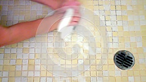 Hand of a woman starts trying to clean dirty shower floor, composed of yellow and white tiles, with a sponge