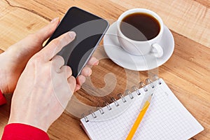 Hand of woman using mobile phone, notepad for writing notes, cup of coffee