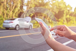 Hand of woman using smartphone and blur of her broken car parking on the road. Contacting car technician or need help concept