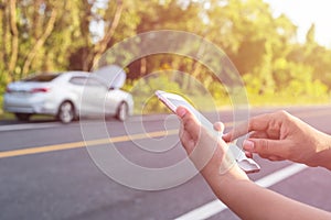 Hand of woman using smartphone and blur of her broken car parking on the road. Contacting car technician or need help concept