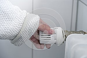 Hand of a woman in warm clothes reduces the heat at the thermostat on the radiator due to the energy crisis with rising prices,