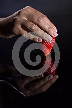 The hand of a woman who takes a strawberry and its reflection with black background