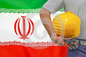 Hand of worker and yellow hard-hat on background flag of Iran