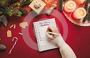 Hand writing in a new year resolution notebook with list of goals and Christmas stuff on red background. Blank space checklist in