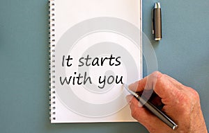 Hand writing `it starts with you` on white note, isolated on blue background