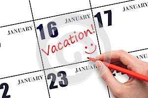 A hand writing a VACATION text and drawing a smiling face on a calendar date 16 January. Vacation planning concept.