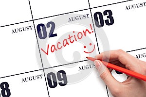 A hand writing a VACATION text and drawing a smiling face on a calendar date 2 August. Vacation planning concept.