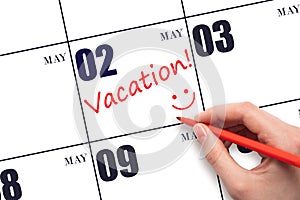 A hand writing a VACATION text and drawing a smiling face on a calendar date 2 May. Vacation planning concept.