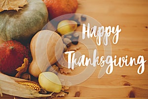 Hand written Happy Thanksgiving text on background of pumpkins, autumn leaves, nuts, harvest vegetables on rustic wooden table.