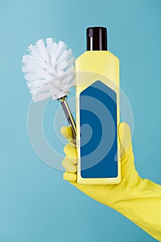 Hand in yellow glove holds bottle of liquid detergent for toilet and brush on blue background.