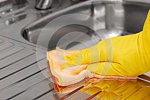 Hand in yellow latex protective glove wiping stainless steel sink with microfiber cloth. Household cleaning tools