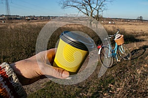 In hand a yellow paper corrugated cup with coffee on a background of nature and a bicycle