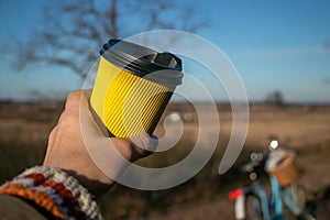 In hand a yellow paper corrugated cup with coffee on a background of nature and a bicycle