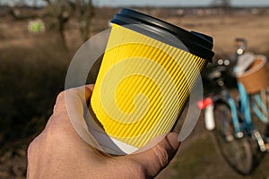 In hand a yellow paper corrugated cup with coffee on a background of nature and a bicycle