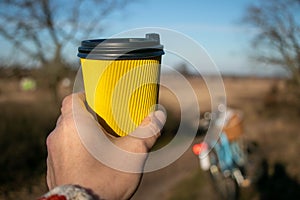 In hand a yellow paper corrugated cup with coffee on a background of nature and a bicycle