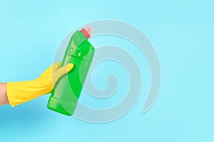 Hand in yellow protective rubber glove holding green spray bottle with detergent against blue background with copy space