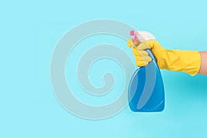 Hand in yellow protective rubber glove holding spray bottle with detergent against blue background with copy space