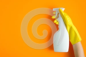 Hand in yellow rubber glove holding plastic spray bottle with cleaning detergent on a orange background