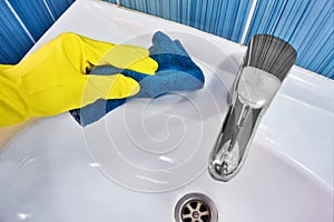 Hand in yellow rubber glove with rag wipes white sink. Cleaning and disinfection with cleaning agent. Clean and shiny