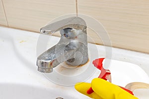 Hand in yellow rubber gloves holding a spray liquid cleaning agent for washing dirty faucet with limescale, calcified water tap