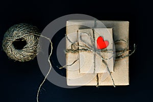 Handcrafted gift box wrapped in Craft paper with red wooden heart, rope and bow on black background. Top view, flat lay, toned