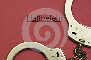 handcuffs next to the german word haftbefehl, translation arrest warrant on a red paper background