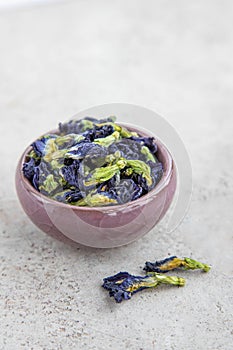 A handful of dried butterfly pea flowers in small cup on a white stone background. Blue flower for tea