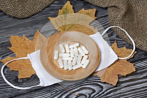 A handful of white pills on a wooden saucer. On a medical mask. Among the dried up maple leaves. On a pine board surface