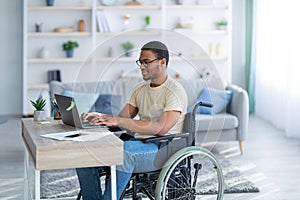 Handicapped black guy in wheelchair working online from home, using laptop, looking for job opportunities on internet