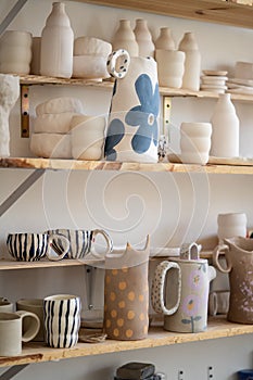 Handmade ceramic cups, jugs, jars and vase on shelves in craft pottery kitchenware store or workshop