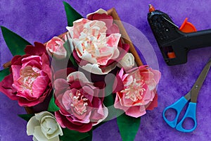 Handmade decorative peonies made from foamiran on wooden panel and glue gun and scissors on purple background