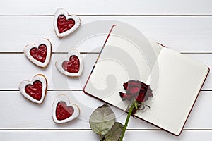 Handmade heart shaped cookies with empty notebook and rose flower on white wooden background for Valentines day.