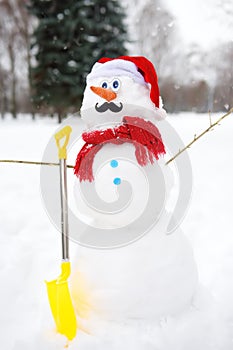 Handmade snowman with a scarf, Santa Claus hat, carrot nose, mustache, hands from branches and shovel in a snowy park