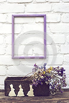 Handmade white bunny on wooden box with multicoloured dry flowers on white bricked wall background with empty violet frame. Easter