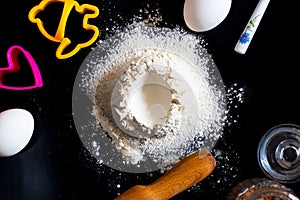 Handrful of sieved flour with egg shape hole in the center and utensils for baking and ingredients around on black background with