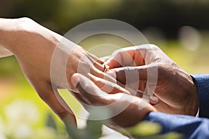 Hands of african american bride and groom placing ring on finger at wedding ceremony in sunny garden