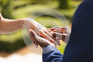 Hands of african american bride and groom placing ring on finger at wedding ceremony in sunny garden